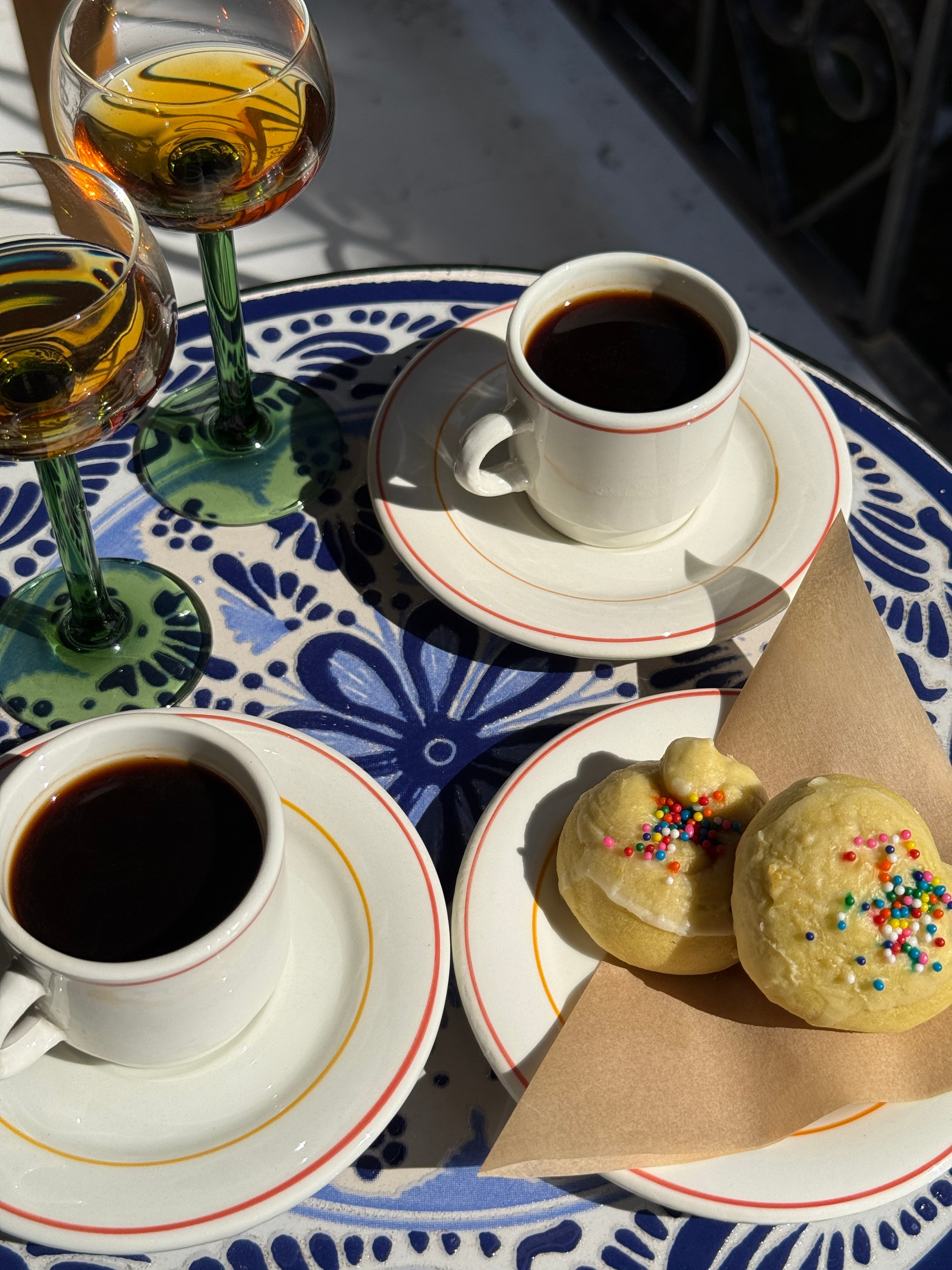 Two cups of coffee, a plate with Italian wedding cookies, and glasses on a decorative tablecloth.