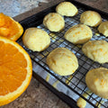 Orange-glazed italian wedding cookies rest on a black wire cooling rack set on a baking sheet, with sliced orange halves visible on the left and a granite countertop underneath.