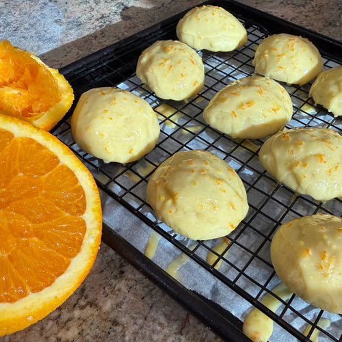 Orange-glazed italian wedding cookies rest on a black wire cooling rack set on a baking sheet, with sliced orange halves visible on the left and a granite countertop underneath.